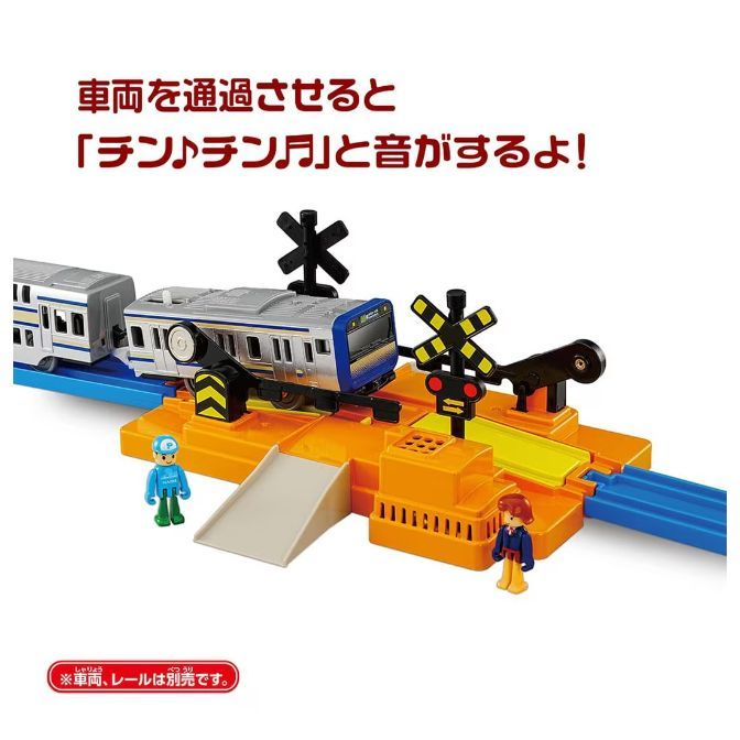  A toy train set with a blue and silver train car passing through a crossing gate. Two toy figures stand nearby, and Japanese text is visible above the train.