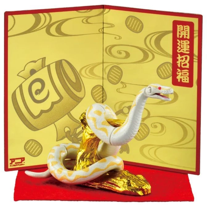 Albino snake toy coiled on gold base, against a decorative backdrop with Japanese text.