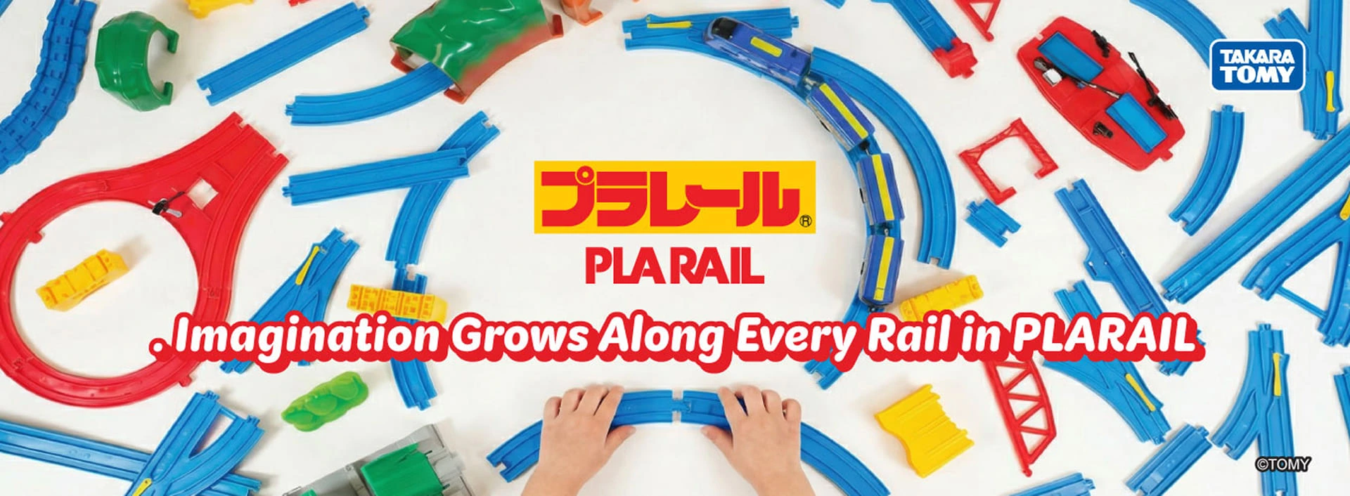 PLARAIL promotional banner showing colorful train tracks and accessories spread across a table, with children’s hands connecting track pieces.