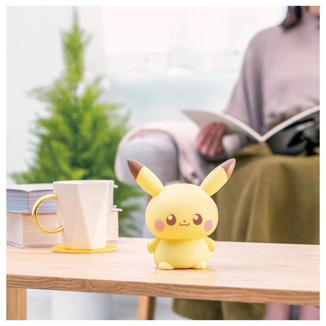  Pikachu toy figure with rosy cheeks on a wooden table, person reading in background.