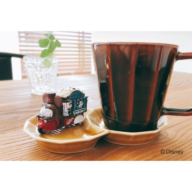  Disney popcorn truck toy on a saucer next to a brown mug, sitting on a wooden table.