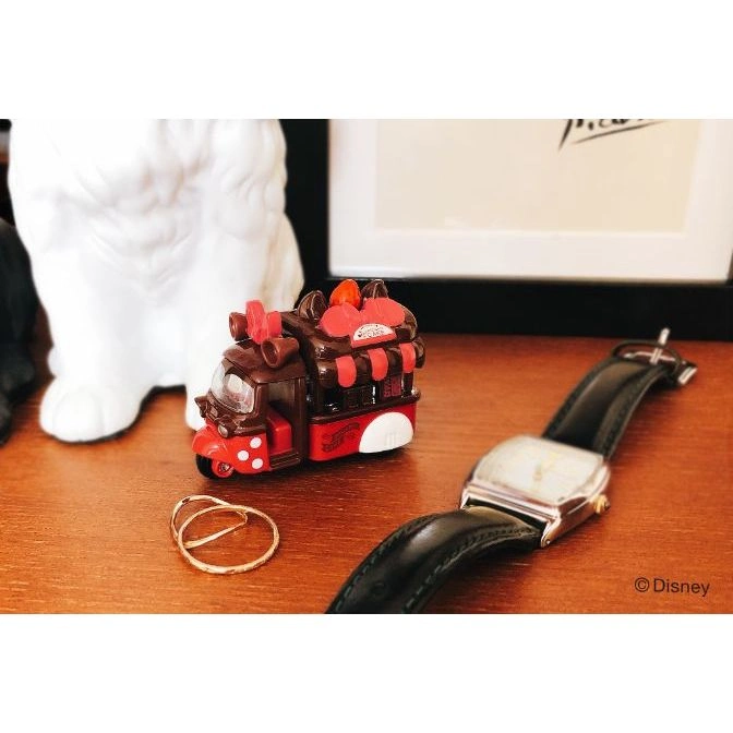  Toy tuk-tuk with a chocolate shop design, gold ring, and a black leather watch on a wooden surface.