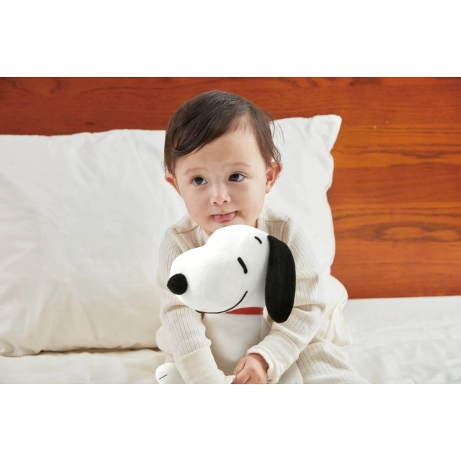  Baby holding a Snoopy plush toy in bed. The toy is white with black ears and a red collar.