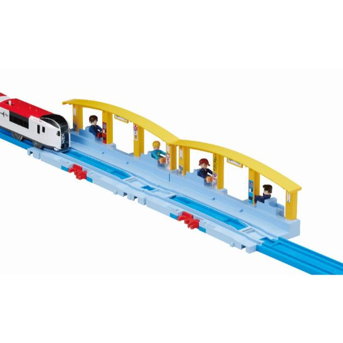  A toy train set with a red and white train approaching a station with yellow awnings and four figures waiting.