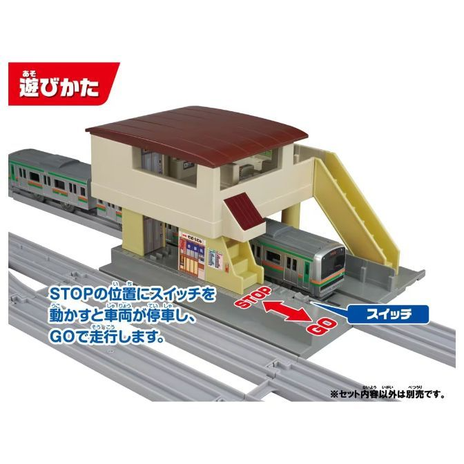  A toy train station with two trains on the tracks. The station is beige with a brown roof and a yellow ramp. There is a sign in Japanese that says "How to play" and instructions on how to use the switch to stop and start the trains.